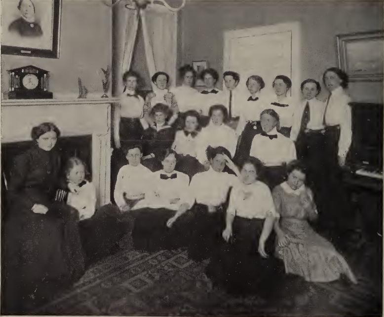 20个人的黑白照片. 所有三行. 大多数人都穿着白色上衣和深色裤子. 在照片的右侧，一个穿着深色连衣裙的女人坐在壁炉前.